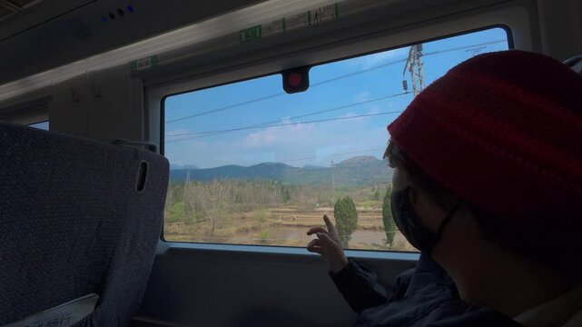 Inside a train carriage, a masked traveler in red points out the window toward fields and mountains under bright skies&mdash;capturing reflective motion, human interaction, and the scenic rhythm of travel
