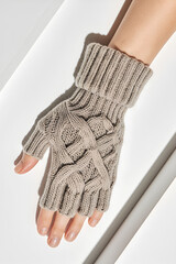 A close-up of a hand wearing a thick, beige knitted glove with an intricate cable-weave pattern, resting on a smooth white surface. The glove features a ribbed cuff that securely wraps the wrist.