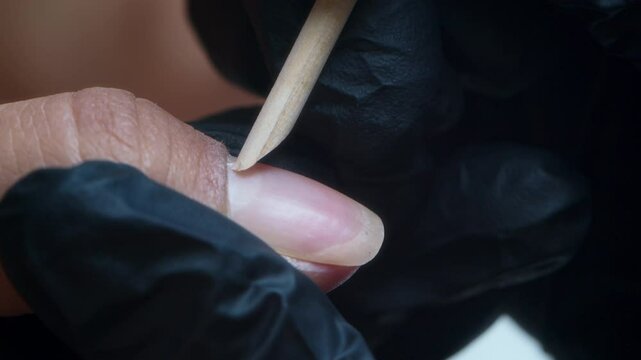 Close-up of professional using wooden cuticle stick to gently push back cuticle during manicure, wearing black gloves for hygiene and precision