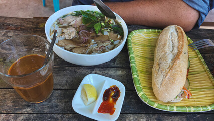 A casual authentic traditional local Vietnamese meal with a bowl of pho bo or beef noodle soup, iced coffee, and a baguette sandwich or banh mi on a rustic wooden table