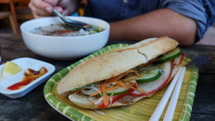 A banh mi sandwich with fresh vegetables with a person eating a bowl of beef noodle soup or pho bo, authentic traditional local Vietnamese cuisine