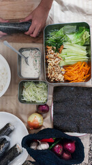 A vertical arrangement of ingredients while hands are rolling a gimbap or seaweed rice roll near vegetables and a sauce bowl
