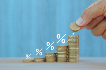  Hand placing a coin on a rising stack with percentage symbols, illustrating increasing interest...