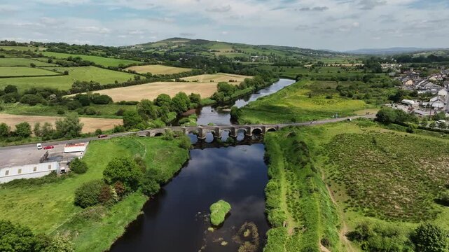 Clady Bridge, County Tyrone, Northern Ireland, June 2023. Drone orbit counter clockwise around arched bridge crossing River Finn with clouds reflecting in calm placid water.