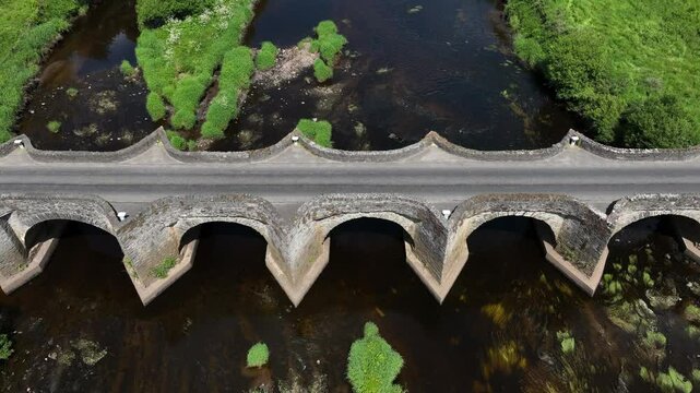 Clady Bridge, County Tyrone, Northern Ireland, June 2023. Drone descends tilting up showcasing arched bridge as cars drive across River Finn.