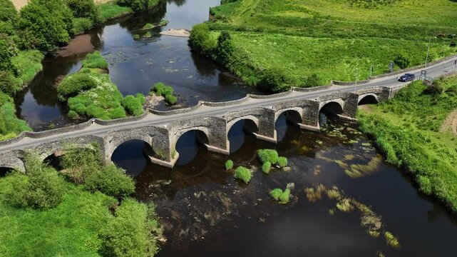 Clady Bridge, County Tyrone, Northern Ireland, June 2023. Drone angled zoom in orbit counterclockwise with arched bridge and symmetrical reflection in water of River Finn.