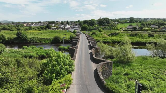 Clady Bridge, County Tyrone, Northern Ireland, June 2023. Drone push in approach over bridge connected towns as line of three cars cross together over the River Finn.