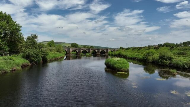 Clady Bridge, County Tyrone, Northern Ireland, June 2023. Drone low approach over River Finn with grassy river bank to arched bridge connecting towns under cloudy sky.