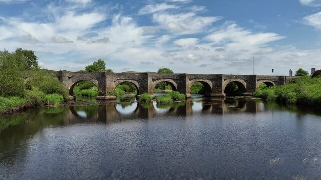 Clady Bridge, County Tyrone, Northern Ireland, June 2023. Drone slow pullback over River Finn with arched bridge centered as cars cross over the pass under a cloudy sky.
