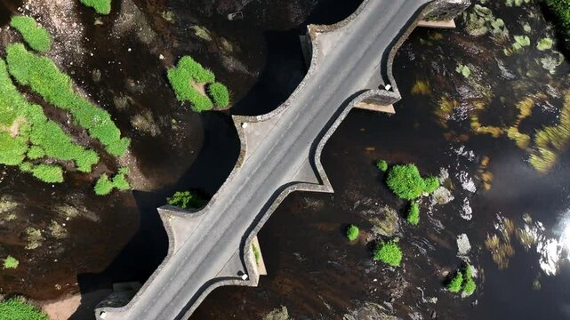 Clady Bridge, County Tyrone, Northern Ireland, June 2023. Drone orbit descends slowly above arched bridge connected land across of River Finn with glistening water.