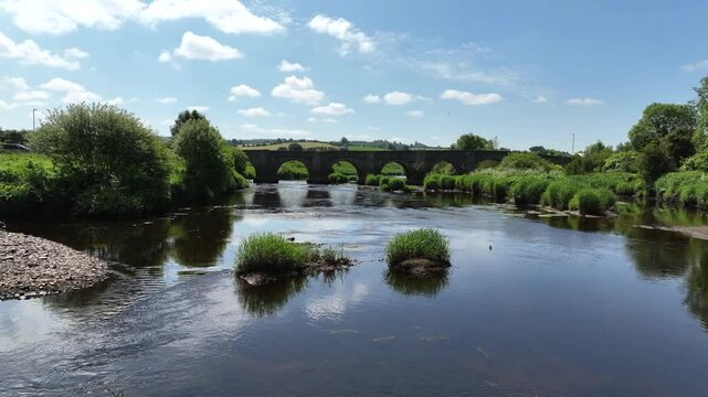 Clady Bridge, County Tyrone, Northern Ireland, June 2023. Drone low approach over shallow River Finn rising ascend over arched bridge as cars cross the water.