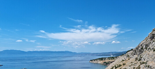 The rocky landscape of Sv. Marko islet, located between the mainland and Krk, is visible under a vast blue sky with distant Rijeka city on the horizon