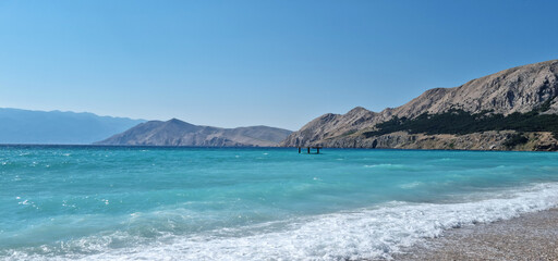 The panoramic view from beach (Vela Plaza) in Baska extends towards the diving board, with the distant island of Prvic visible across the vibrant turquoise sea and the rugged coastline
