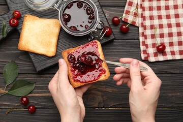 Woman making toast with tasty cherry jam at wooden table, top view