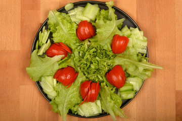 A top-down view displays a vibrant salad with fresh lettuce and cucumber slices arranged around artfully cut tomato pieces on a dark plate, all on a wooden surface