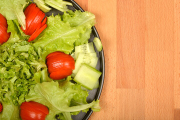 A top-down view displays a vibrant salad with fresh lettuce and cucumber slices arranged around artfully cut tomato pieces on a dark plate, all on a wooden surface
