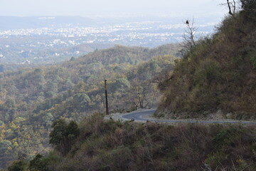 MUSSOORIE, INDIA – March 24,2024: Curving hillside mountain road with Dehradun city visible in the valley below, forested slopes, and misty Himalayan foothills.