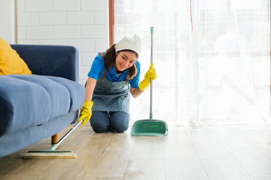 Smiling female cleaner kneels on the floor while sweeping dust and debris from under a blue sofa into a dustpan, showcasing a thorough home cleaning service in a bright, tidy living space. - Powered by Adobe