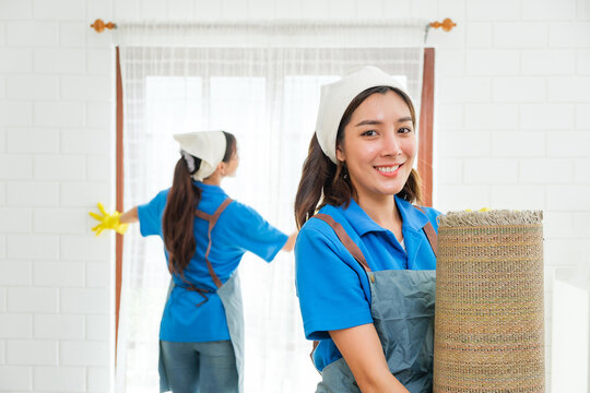 Two professional female cleaners in uniform working in a bright modern home—one smiling while holding a rolled rug, the other cleaning windows—showcasing expert home cleaning service efficiency.