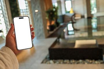 Hand holding smartphone with blank white screen in modern spa interior with indoor pool, plants,...