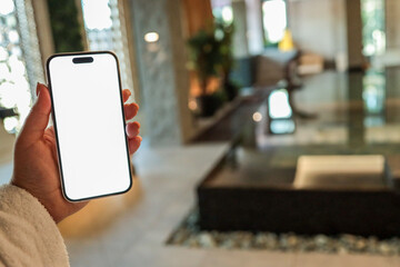 Hand holding smartphone with blank white screen in modern spa interior with indoor pool, plants,...