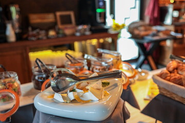 Hotel breakfast buffet with plate of sliced soft cheese, orange slices, tongs, and jars of jam or yogurt. Fresh and elegant food presentation in warm, cozy dining area