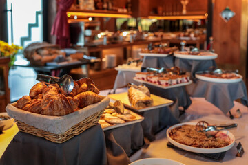 Hotel breakfast buffet with basket of croissants, assorted pastries, cakes, and desserts. Elegant self-service table with tongs, tablecloths, and warm lighting in a cozy dining area