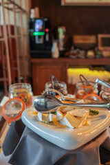 Hotel breakfast buffet with plate of sliced soft cheese, orange slices, tongs, and jars of jam or yogurt. Fresh and elegant food presentation in warm, cozy dining area