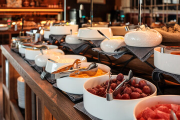 Buffet counter with bowls of fresh fruit, cheese, and jams. Close-up of grapes, melon, and watermelon pieces in white bowls with serving tongs. Elegant breakfast setup in a hotel restaurant