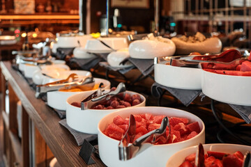 Buffet counter with bowls of fresh fruit, cheese, and jams. Close-up of grapes, melon, and watermelon pieces in white bowls with serving tongs. Elegant breakfast setup in a hotel restaurant