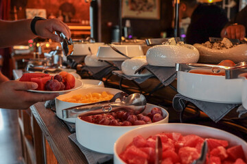 Person serving fresh fruit at a breakfast buffet. Plates filled with grapes, watermelon, oranges, apricots, and strawberries. Elegant setting with bowls and tongs on wooden counter