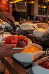 Person serving fresh fruit at a breakfast buffet. Plates filled with grapes, watermelon, oranges, apricots, and strawberries. Elegant setting with bowls and tongs on wooden counter