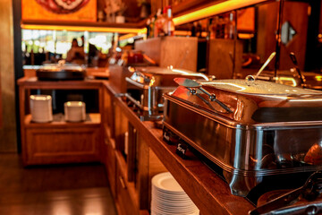 Close-up of metal chafing dishes on a wooden buffet counter in a restaurant. Warm lighting, modern catering equipment, ready for serving hot food at an event or breakfast