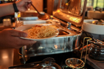 Close-up of person serving spaghetti with tomato sauce at buffet. Warm restaurant lighting,...
