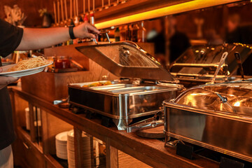 Buffet counter with stainless steel food warmers and a person holding a plate of spaghetti. Warm...