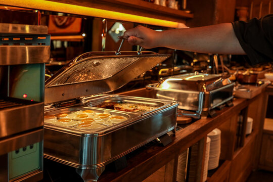 Buffet station with stainless steel food warmers, open lid revealing fried eggs and other breakfast dishes. Hand lifting lid, warm lighting, catering or hotel breakfast setting