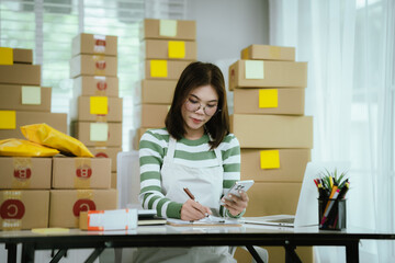 A young Asian woman entrepreneur in a striped shirt and apron works from her home office, managing e-commerce inventory, packaging orders, and organizing shipping for online customers.