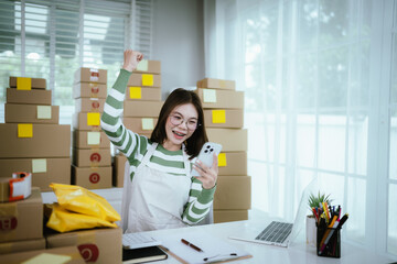 A young Asian woman entrepreneur in a striped shirt and apron works from her home office, managing e-commerce inventory, packaging orders, and organizing shipping for online customers.