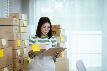 A young Asian woman entrepreneur in a striped shirt and apron works from her home office, managing e-commerce inventory, packaging orders, and organizing shipping for online customers.