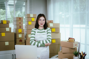 A young woman entrepreneur manages her online business from home, surrounded by parcel boxes, using...