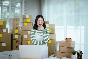 A young woman entrepreneur manages her online business from home, surrounded by parcel boxes, using a laptop, calculator, and notepad to handle shipping and inventory.
