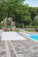 Elegant outdoor wedding setup with floral arch and poolside chairs near a lush garden
