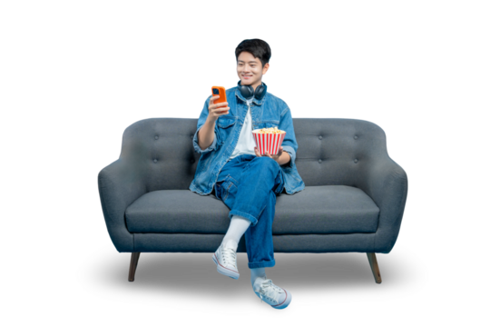 PNG,A young Asian man relaxes on a sofa with popcorn, smiling as he browses or texts on his smartphone.
