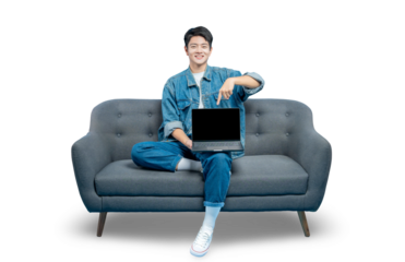 A smiling young man sits on a sofa and points at a laptop with a blank screen, recommending a website or online service.PNG
