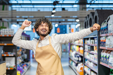 A joyful man with curly hair in a grocery store, wearing an apron, arms raised in celebration.