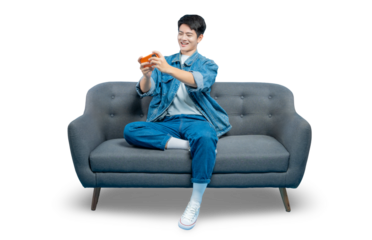 A happy young man in a denim jacket plays a game on his smartphone while relaxing on a sofa.PNG