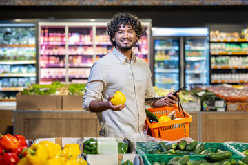 A smiling man holding a yellow bell pepper while shopping in a supermarket. He is looking at the camera, carrying a basket of groceries.