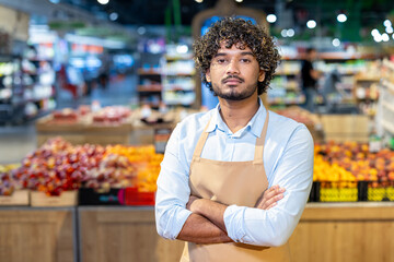 A man with curly hair stands in a supermarket, arms crossed, wearing an apron. Fruits are visible...