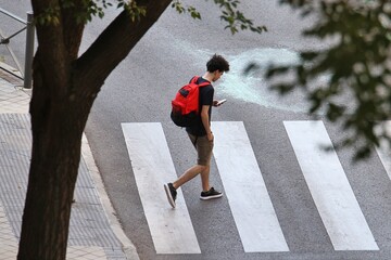 Chico joven mirando el teléfono móvil con mochila roja cruzando la calle por un paso de peatones