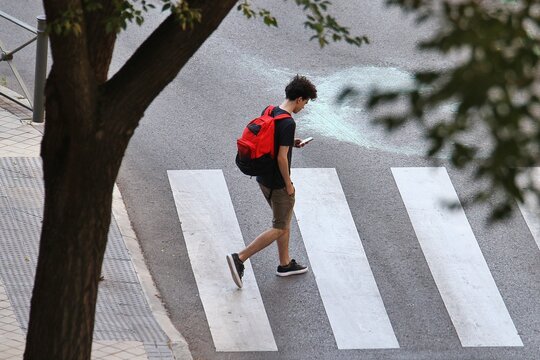 Chico joven mirando el tel&eacute;fono m&oacute;vil con mochila roja cruzando la calle por un paso de peatones